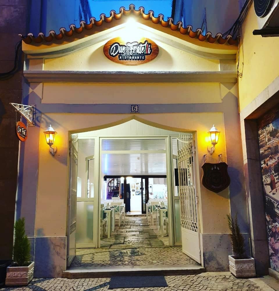 Entrada do Due Fratelli Ristorante à noite, com letreiro de madeira e portões de ferro abertos